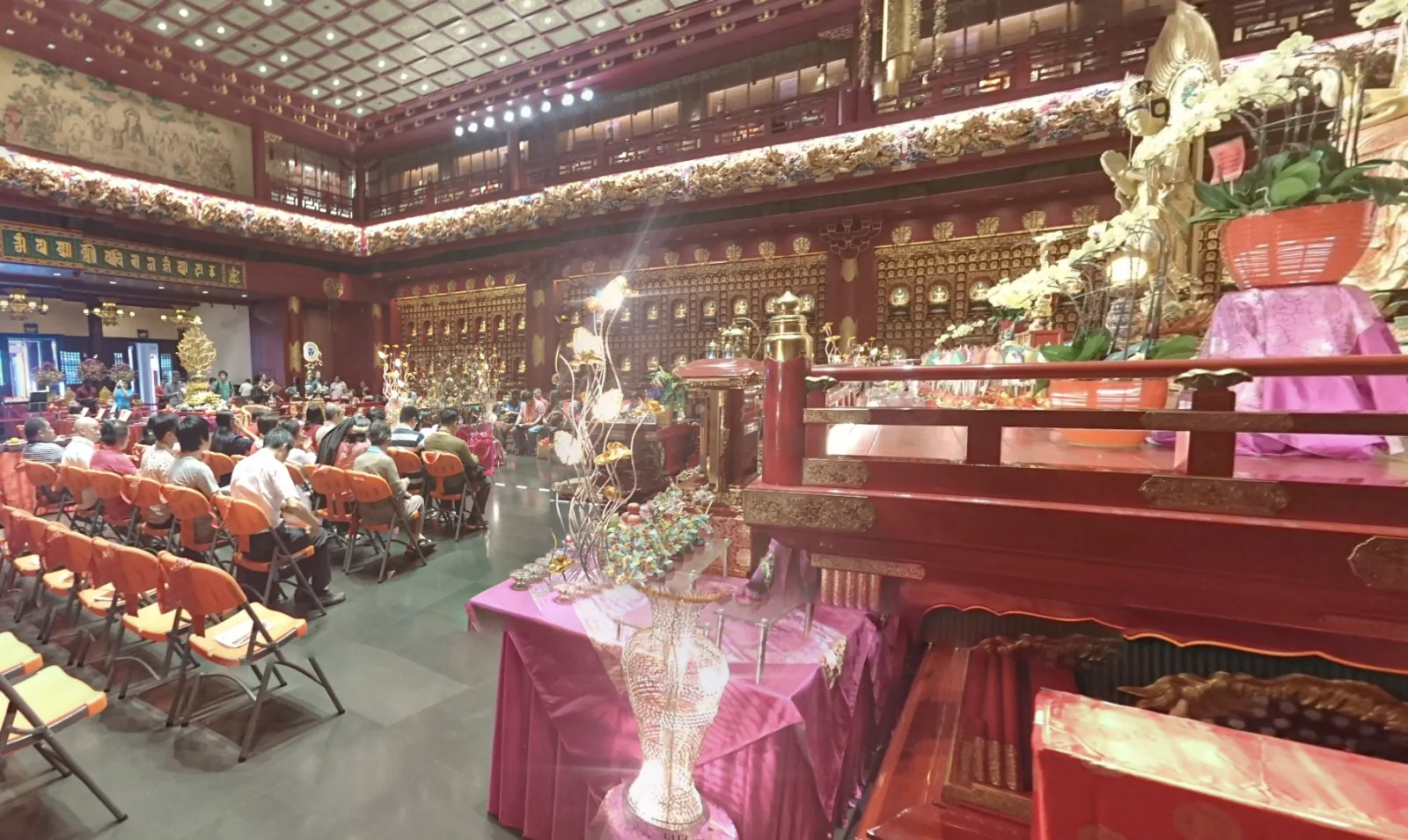 Inside the Buddha Tooth Relic Temple, people sit facing a shrine amid red columns and offerings